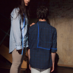 A man and woman are still. He is knelt wearing a dark blue shirt and light trousers. The woman stands wearing the same colours.