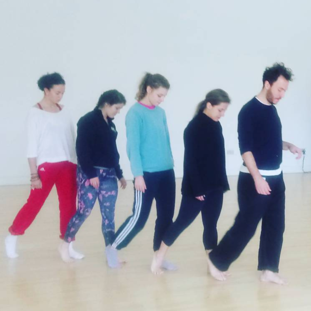 Five people wearing different coloured clothing walk in a line. They are looking at the floor as they move through the dance studio.