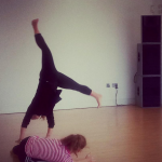 Two women dance together in a studio. One wearing black is in mid cartwheel, the other in a pink top and dark trousers is crouching on the floor.