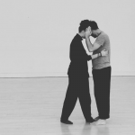 A black and white image of two dancers. They are hiding their face away from the camera as they lean towards each other.