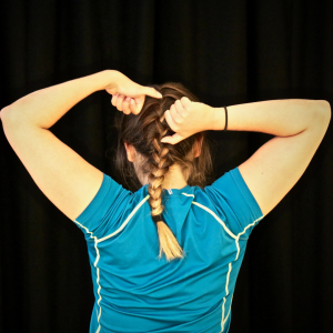 A woman has her back to the camera. Her arms are lifted behind her head and her thumbs are out. Her brown hair is plaited and she is wearing a blue top.