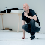 Choreographer Wayne McGregor crouches in his white studio as he observes the action.