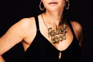 A photo of a white woman in a black top wearing a gold necklace that says Rebel Boob