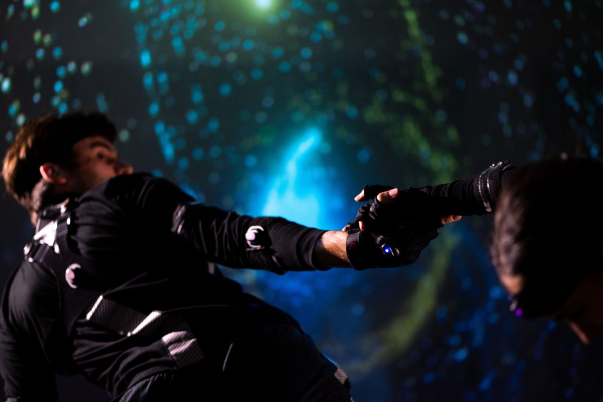 A male dancer all in black and wearing a motion capture suit looks behind him, towards his arm that is stretched out to hold the hand of another dancer. The background is a projection of halos on a screen.