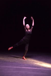 A female dancer in black holding their hand out and stretching into the air with a full leg spread
