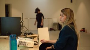 Image of creative in VR headset and another artist sitting at a desk looking at a computer.