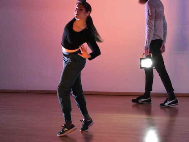 Two dancers, one in black top and the other in a grey top practicing a dance routine together on stage