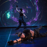 Two dancers are wearing all black and motion capture suits. They dance in front of a pretty background of pink and blue swirls. One is facing the back, her arms outstretched. The other is on the floor.