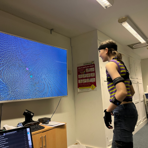 A young person is looking at her and her partners avatar on the tv screen as she is wearing a mocap suit.