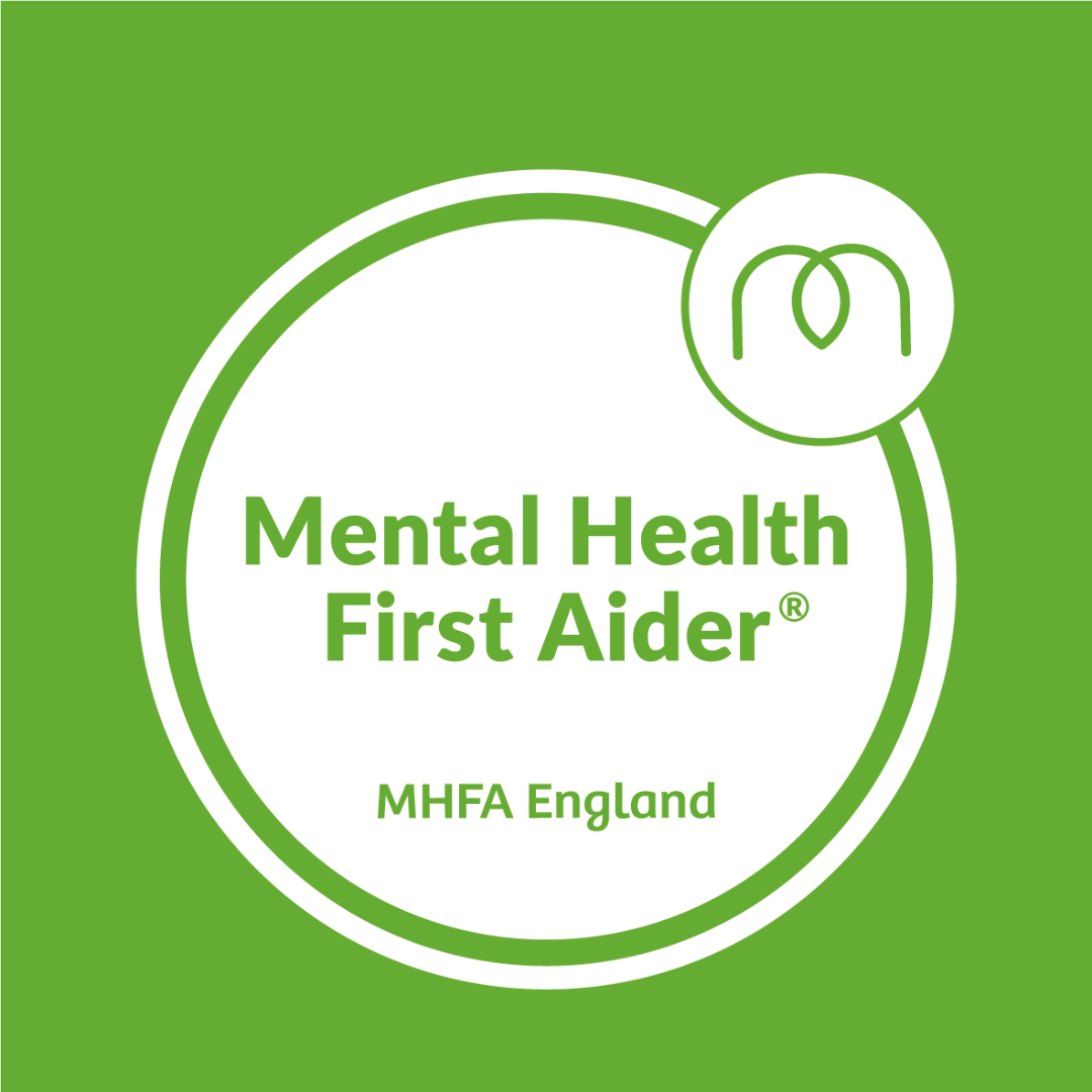 A Mental Health First Aider Badge by MHFA England. The colours in the badge are green and white, the text is in a circle and the MHFA England logo (which looks like the letter 'M' in the top right of the circle.