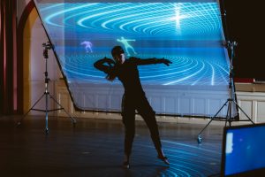 A woman dances in front of a screen with projection on it.