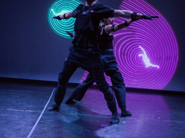 Two people dressed in black performing with neon lights in the background and a circular design on the wall behind them