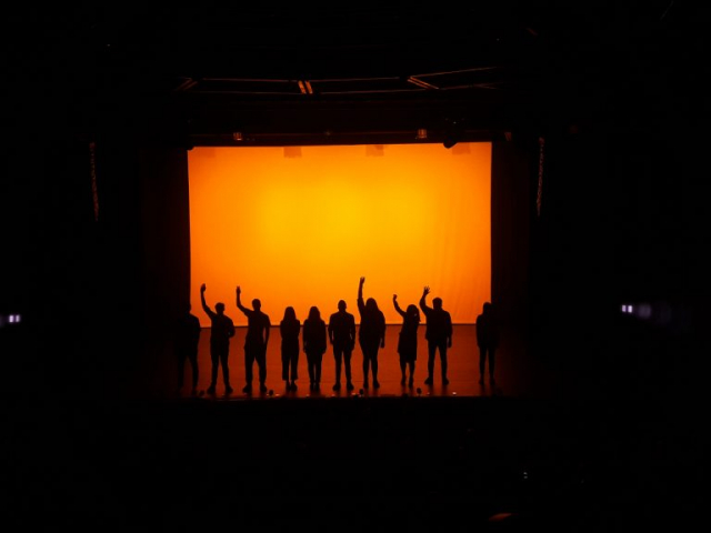 A group of people standing on top of a stage with an orange lit backdrop.